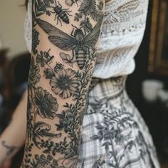 a woman's arm covered in tattoos with flowers and bees on it, while she is wearing a white top