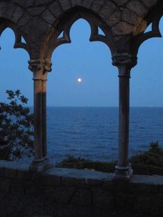 the moon is setting over the ocean from an old building with arches and stone pillars