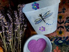 a purple heart in a bowl next to lavender flowers and a pot with a dragonfly painted on it