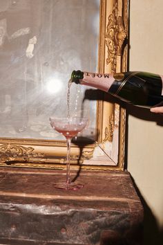 a person pouring wine into a glass in front of a framed painting on the wall