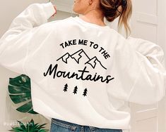 a woman wearing a white sweatshirt with the words take me to the mountains printed on it