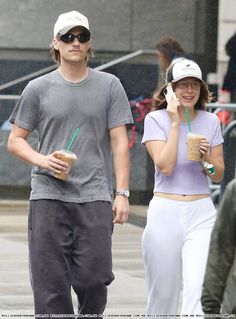 a man and woman walking down the street while talking on their cell phones with drinks in each hand