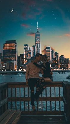 a man and woman are kissing on a pier in front of the city at night