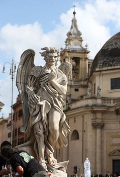 the statue of an angel in front of a building with people walking around and looking at it