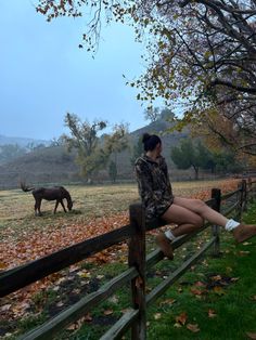 a woman sitting on a fence looking at an animal in the distance with leaves all around her