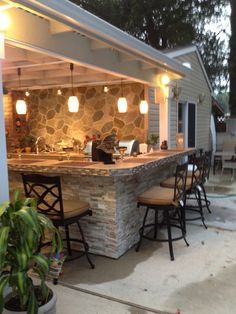 an outdoor kitchen is lit up at night with lights on the counter and chairs around it