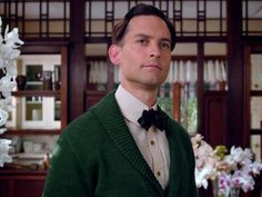 a man wearing a green sweater and bow tie in a living room with white flowers