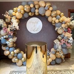 a large balloon arch with flowers and balloons on the wall next to a gold stool