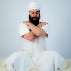 a man with a beard is sitting on the floor in white clothing and has his arms crossed