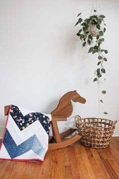 a wooden rocking horse next to a basket with a quilt on it and a potted plant in the corner