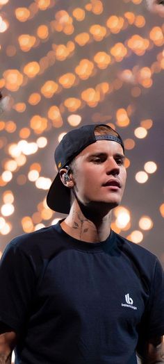 a man with tattoos wearing a baseball cap and looking off to the side while standing in front of lights