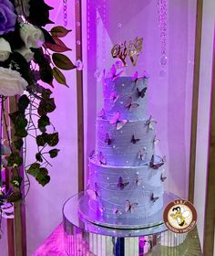 a three tiered cake with butterflies on the top and purple lighting behind it, surrounded by flowers