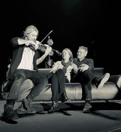 three people are sitting on a couch playing the violin while another person is standing next to them