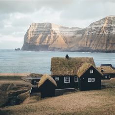 two black houses with grass roofs near the ocean and mountains in the background on a cloudy day