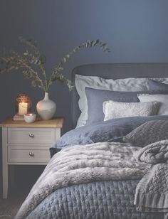 a bedroom with dark blue walls and grey bedding in the foreground is a white table with two vases on it