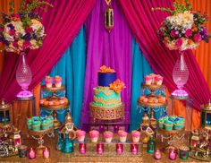 a table topped with lots of cakes and cupcakes next to colorful drapes