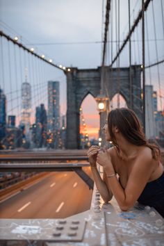 a woman sitting on a bench in front of the brooklyn bridge at sunset with her arms crossed