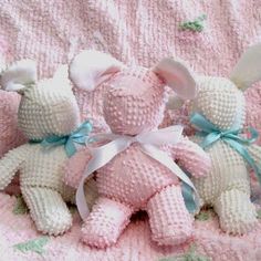 three small knitted teddy bears sitting on a pink blanket with blue ribbons and bows