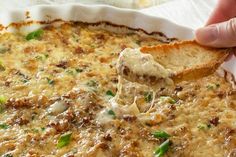 a hand holding a piece of bread over a casserole filled with cheese and meat