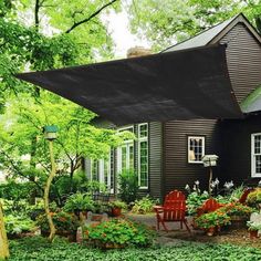 a black house in the woods with lots of plants and flowers around it, along with two red chairs