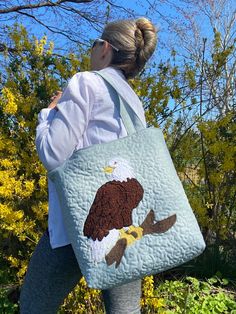 "This handmade bag has a beautiful bold Bald Eagle perched on a branch. This bag was made in mind for all of those eagle nest followers. So many famous nest cams are watched today. I have followed several nest over the years and have been inspired to make these bags. This bag makes a great gift and will last for many years.  The bag is approximately 13\"x 14\". It is extremely durable and lined. It has 3 inside pockets. It also has a magnetic clasp that opens and closes easily. The bag has nice long padded handles approximately 27 \" long. The front of the bag is quilted. Each bag I make is one of a kind because it is done freehand by me. The bags are also hand pieced and stitched in our smoke free home. Free shipping included with this purchase." Quilted Pouch Bag As Gift, Quilted Pouch Bag For Gift, Quilted Rectangular Bag For Gifts, Quilted Rectangular Bag For Gift, Quilted Rectangular Bag As Gift, Rectangular Quilted Bag For Gifts, Rectangular Quilted Bag As Gift, Eagle Quilt, Eagle Nest