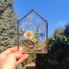 a hand holding up a small glass house with flowers in the front and butterflies on the back