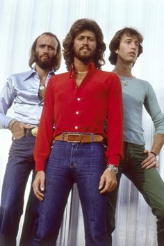 three men standing next to each other in front of a metal wall wearing red shirts and blue jeans