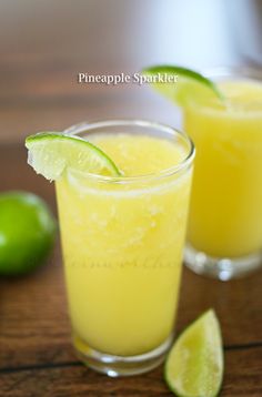 two glasses filled with drinks sitting on top of a wooden table next to limes