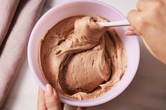 a person holding a spoon in a bowl filled with chocolate frosting on top of a table