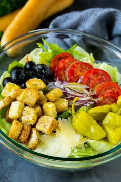 a salad with croutons, olives, tomatoes and lettuce in a glass bowl