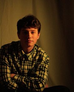 a young man sitting in front of a white wall wearing a plaid shirt and black pants