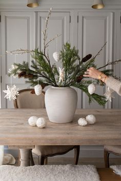 a woman arranging flowers in a white vase on a wooden table next to a chair