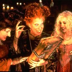 three women in costume looking at something on a table with candles behind them and one holding a book