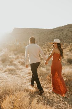 a man and woman holding hands while walking through the desert with mountains in the background