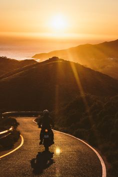 a motorcyclist rides down a winding road at sunset with the sun setting