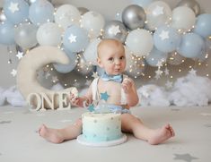 a baby sitting on the floor with a cake in front of it and balloons behind him