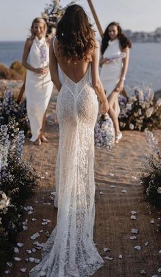 three women in white dresses walking down a path next to the ocean with flowers all around them