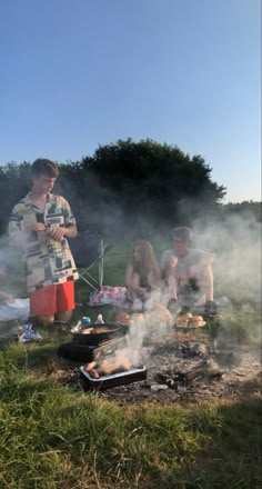 a group of people sitting around a campfire with food cooking on the fire pit