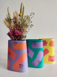 three colorful vases with flowers in them on a white surface, one is painted bright and the other has an abstract design