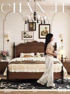 a woman walking across a bedroom next to a bed in front of a chandelier