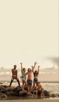 four people standing on rocks at the beach with their arms in the air and one person jumping up to catch a frisbee