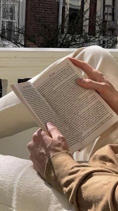 a woman sitting in a chair reading a book on top of a white couch next to a window