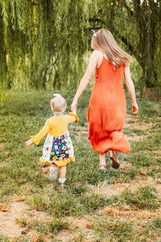 a mother and her toddler walking in the grass under a willow tree holding hands