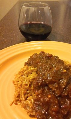 a yellow plate topped with meat and rice next to a glass of wine on a table