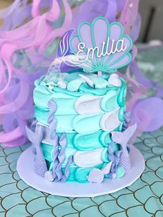 a blue and purple cake with sea shells on it sitting on a table next to streamers