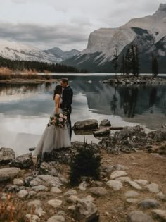 a bride and groom standing on the shore of a mountain lake