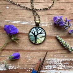 a necklace with an image of a tree on it next to purple flowers and paintbrushes