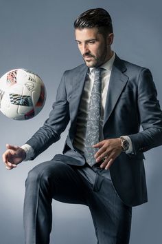 a man in a suit and tie holding a soccer ball with his right hand while sitting on a chair