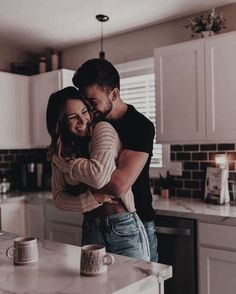 a man and woman hug in the kitchen while standing next to an island with two mugs on it