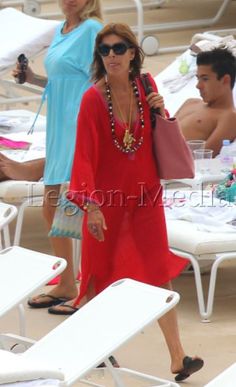 a woman in a red dress and sunglasses walking on the beach with her handbag